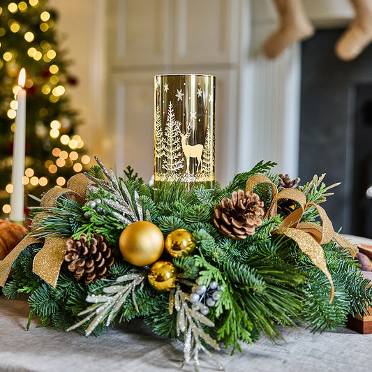 A festive Christmas centerpiece with a gold cylindrical candle holder featuring a deer and tree design, surrounded by green pine branches, gold ornaments, pinecones, and a gold ribbon, set on a table in a softly blurred holiday interior.