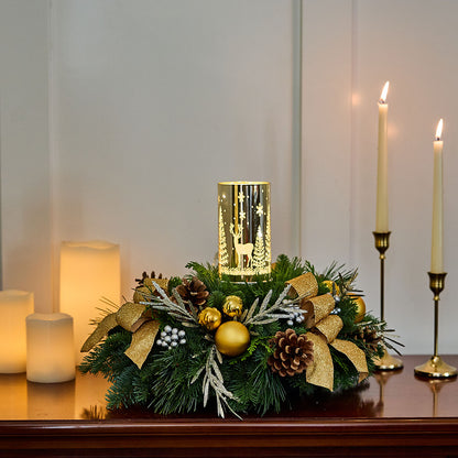 A festive Christmas wreath centerpiece on a dark wooden mantel, decorated with green pine branches, gold ribbons, pinecones, white berries, and gold ornaments, with a glowing gold cylindrical lantern mounted on top. Lit pillar candles in brass holders stand in the background against a soft gray wall.