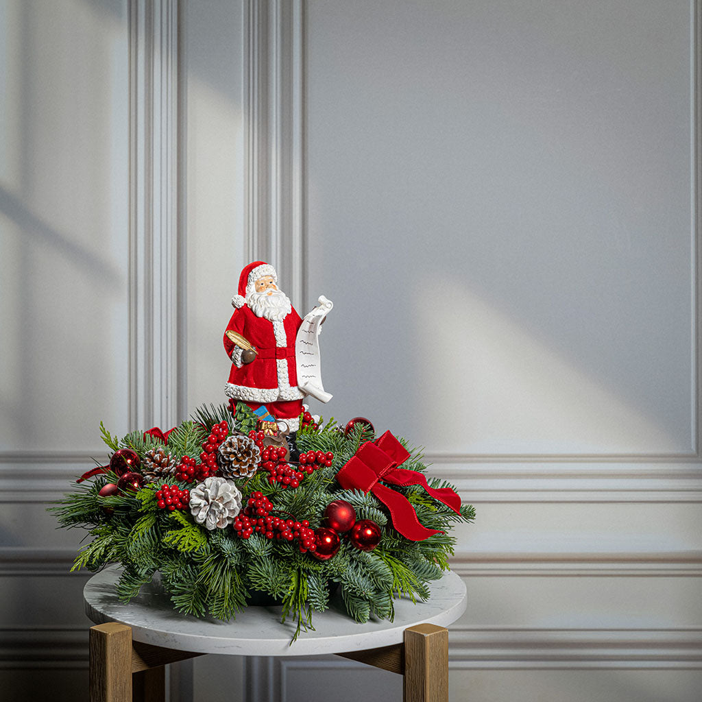 A festive Christmas centerpiece decoration displayed on a small round table against a light gray paneled wall. The centerpiece is made of green pine branches with red berries, pinecones, ornaments, and a large red bow, topped with a Santa Claus figurine holding a scroll.