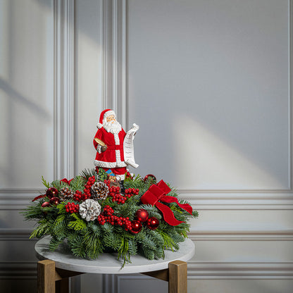 A festive Christmas centerpiece decoration displayed on a small round table against a light gray paneled wall. The centerpiece is made of green pine branches with red berries, pinecones, ornaments, and a large red bow, topped with a Santa Claus figurine holding a scroll.