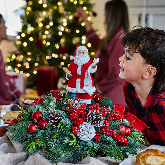 A festive Christmas centerpiece with a Santa Claus figurine, green pine branches, red ornaments, and pinecones, shown on a table in a decorated living room with a child looking at it.