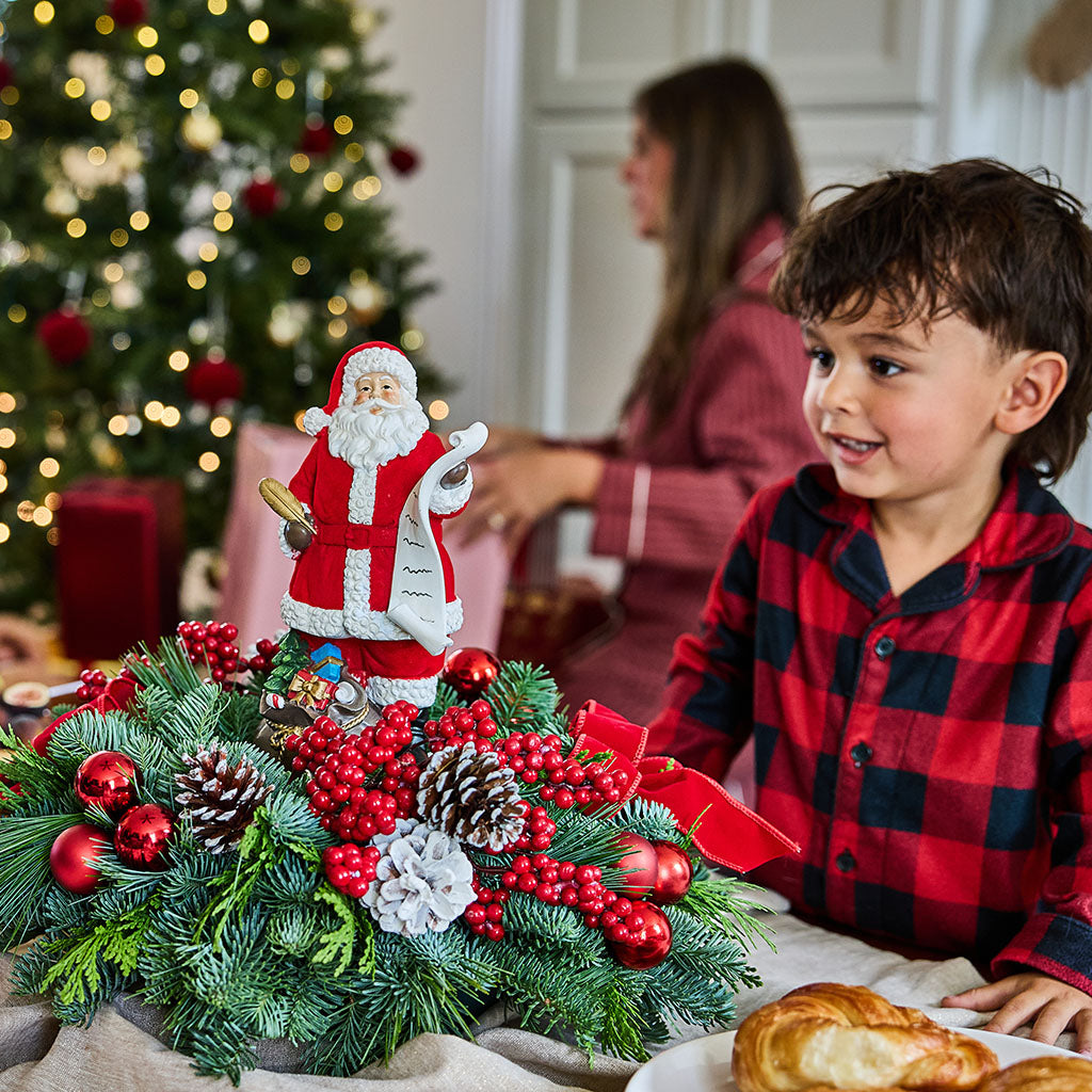A young boy in a red and black plaid pajama shirt smiles while sitting at a table with a festive Christmas centerpiece, a Santa Claus figurine, and a blurred Christmas tree in the background.
