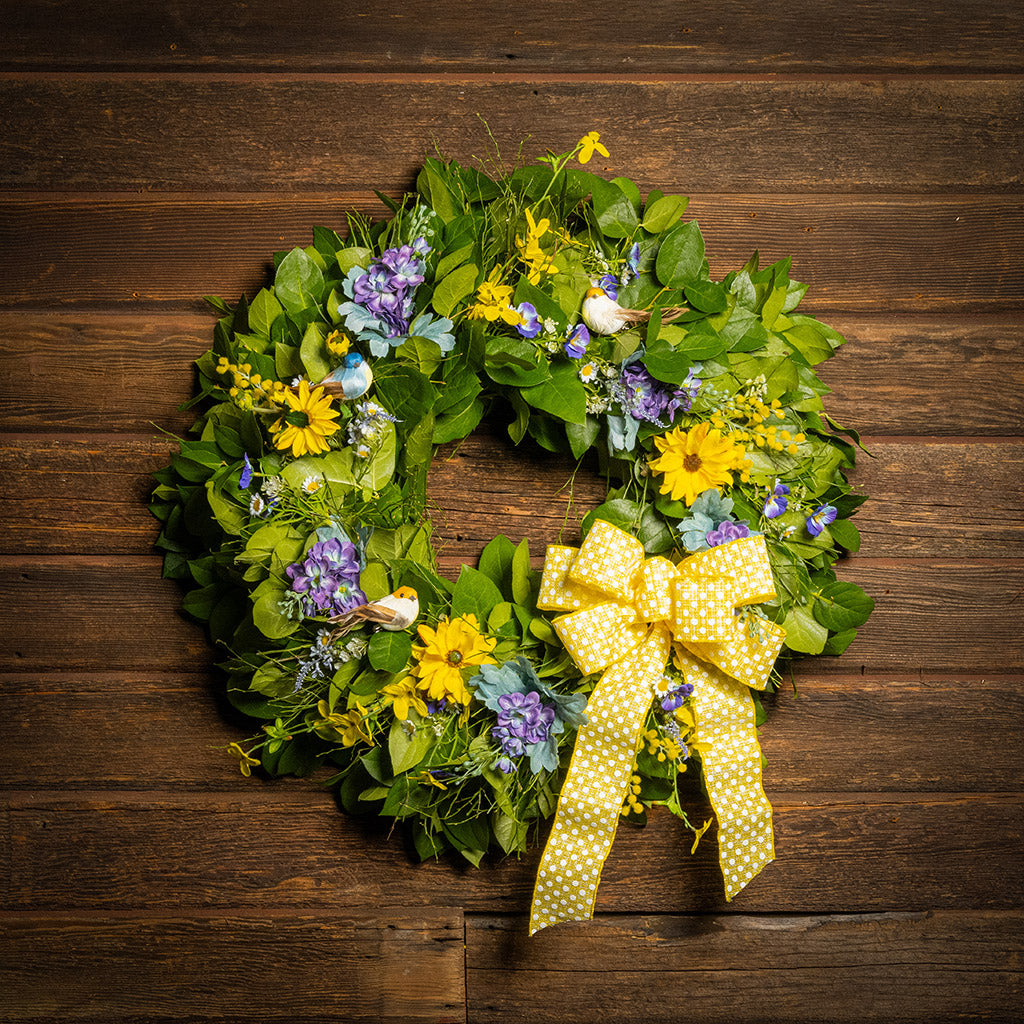 Fresh salal and sweet huckleberry with faux lavender pansies, faux yellow forsythia flowers, faux purple delphinium flowers, faux yellow daisies, and three birds with a yellow polka-dot bow