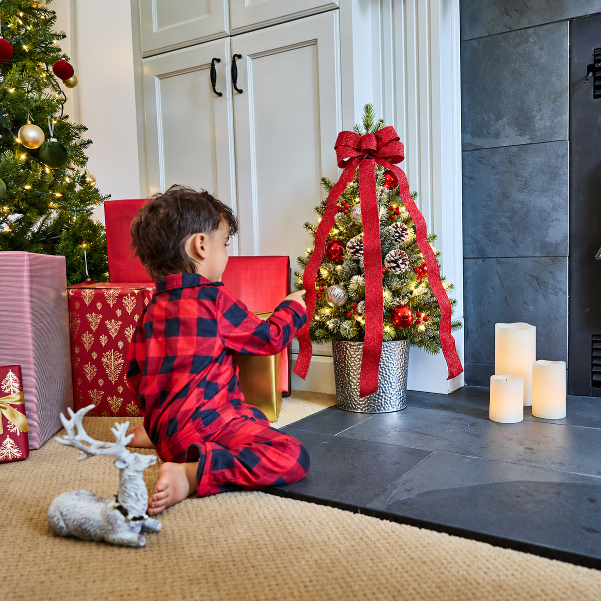 A child wearing red and black buffalo plaid pajamas sits on a beige rug in a holiday living room, looking at a small decorated Christmas tree with a large red ribbon. Gift boxes, a lit Christmas tree, candles, and a fireplace are visible in the background.