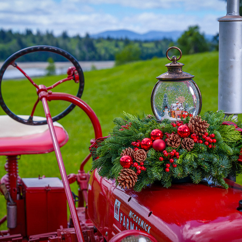 An evergreen arrangement of noble fir, cedar, and pine with red ball ornaments, faux red berries, pinecones, plaid bow tucks, and a farm-scene water globe sitting on a red tractor with a green grass background.