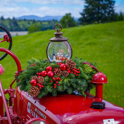 An evergreen arrangement of noble fir, cedar, and pine with red ball ornaments, faux red berries, pinecones, plaid bow tucks, and a farm-scene water globe sitting on a red tractor with a green grass background.