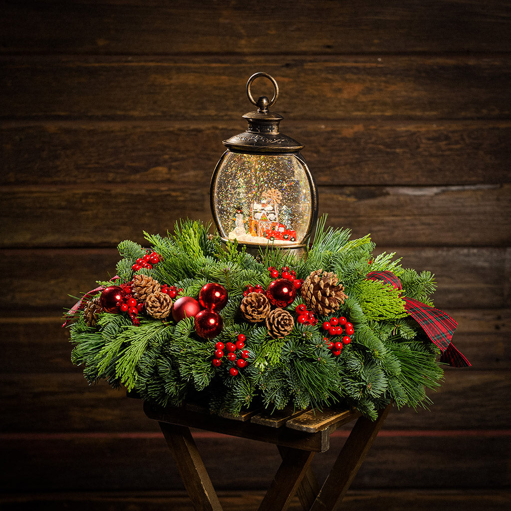 An evergreen arrangement of noble fir, cedar, and pine with red ball ornaments, faux red berries, pinecones, plaid bow tucks, and a farm-scene water globe with a dark wood background.