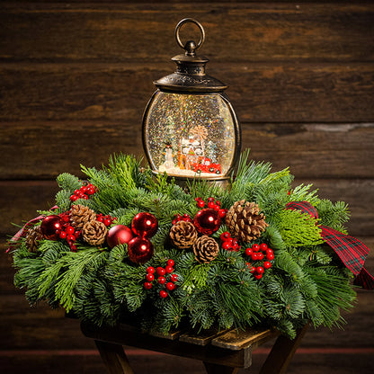 An evergreen arrangement of noble fir, cedar, and pine with red ball ornaments, faux red berries, pinecones, plaid bow tucks, and a farm-scene water globe with a dark wood background.