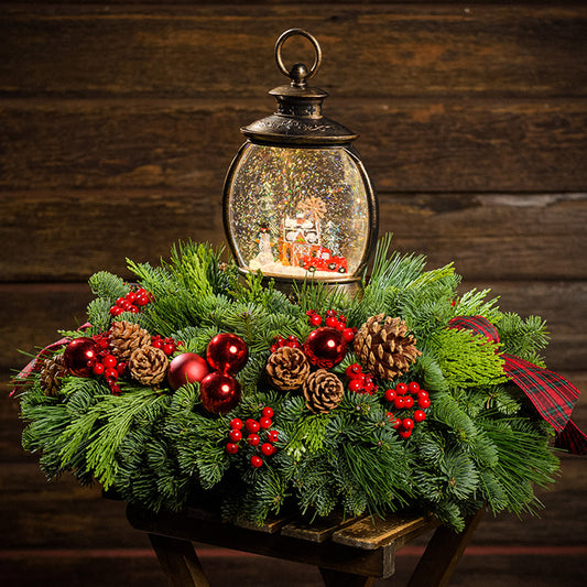 An evergreen arrangement of noble fir, cedar, and pine with red ball ornaments, faux red berries, pinecones, plaid bow tucks, and a farm-scene water globe with a dark wood background.