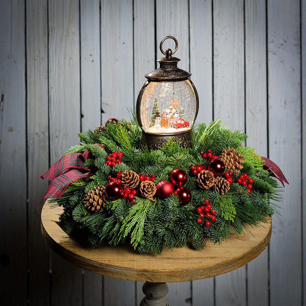 An evergreen arrangement of noble fir, cedar, and pine with red ball ornaments, faux red berries, pinecones, plaid bow tucks, and a farm-scene water globe sitting on a table with a white wood background.