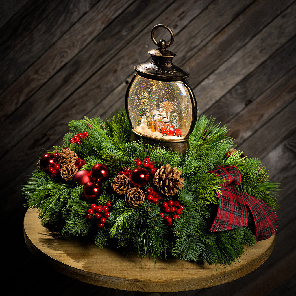 An evergreen arrangement of noble fir, cedar, and pine with red ball ornaments, faux red berries, pinecones, plaid bow tucks, and a farm-scene water globe sitting on a table with a wood background.