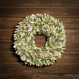 A dried wreath made of natural green fern, integrifolia leaves, bleached phalaris, English lavender, and white daisies hung on a dark wood background.