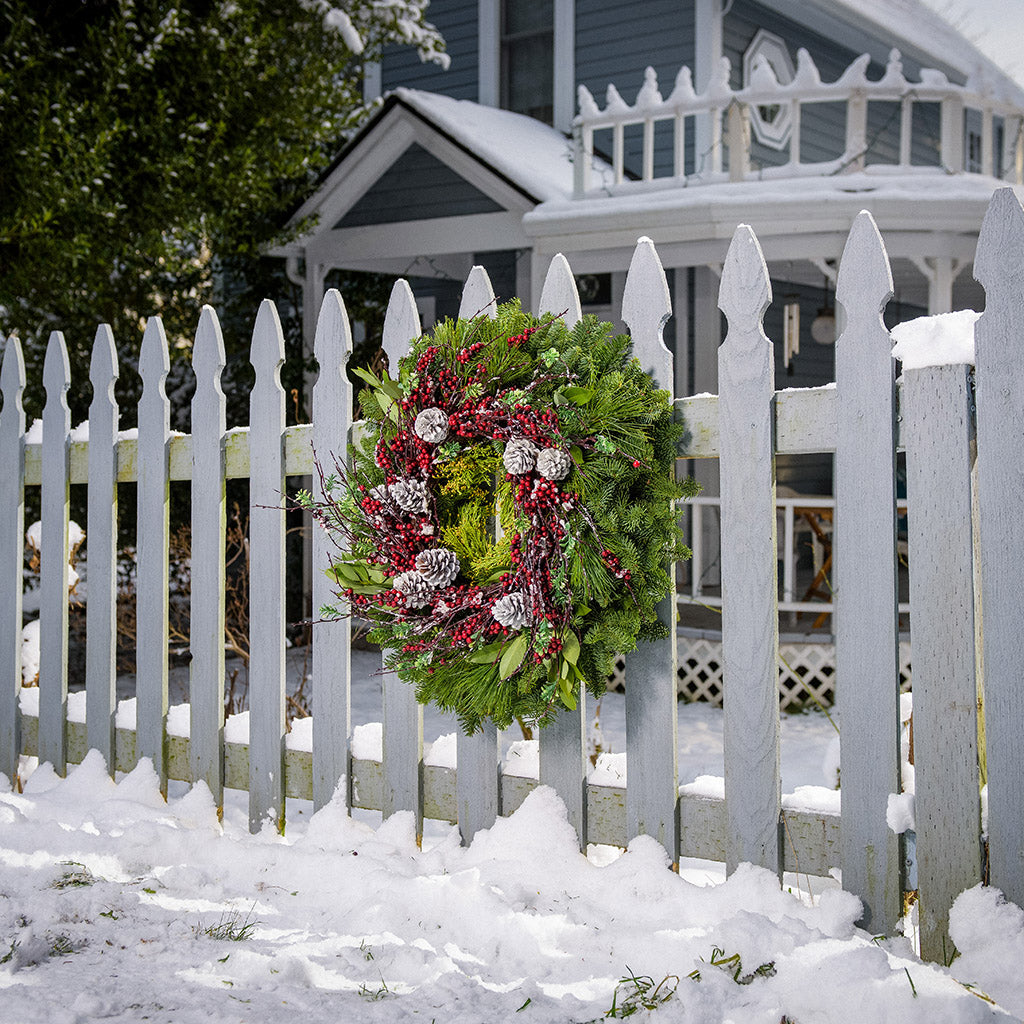 Christmas wreath made of noble fir, cedar, white pine, and bay leaves with 9 frosted Australian pine cones, and a ring of frosted branches with faux burgundy berries hanging on a white fence