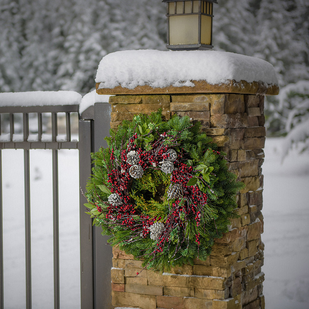 Christmas wreath made of noble fir, cedar, white pine, and bay leaves with 9 frosted Australian pine cones, and a ring of frosted branches with faux burgundy berries hanging on a white fence