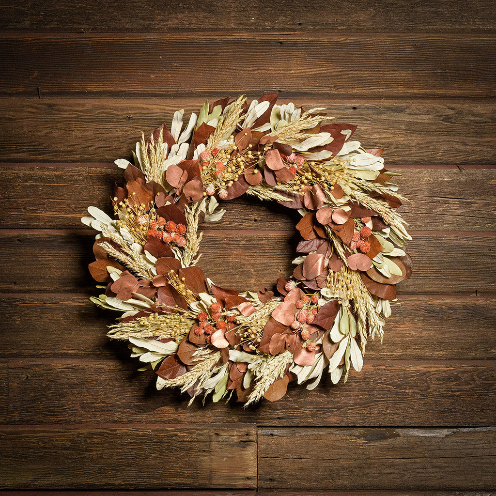 A dried wreath made of rich preserved deep burnt orange salal leaves, copper silver dollar eucalyptus leaves, flax pods, sage green integrifolia leaves, and red-orange globe amaranthus with long wispy sudan grass accents hung on a dark wood background.
