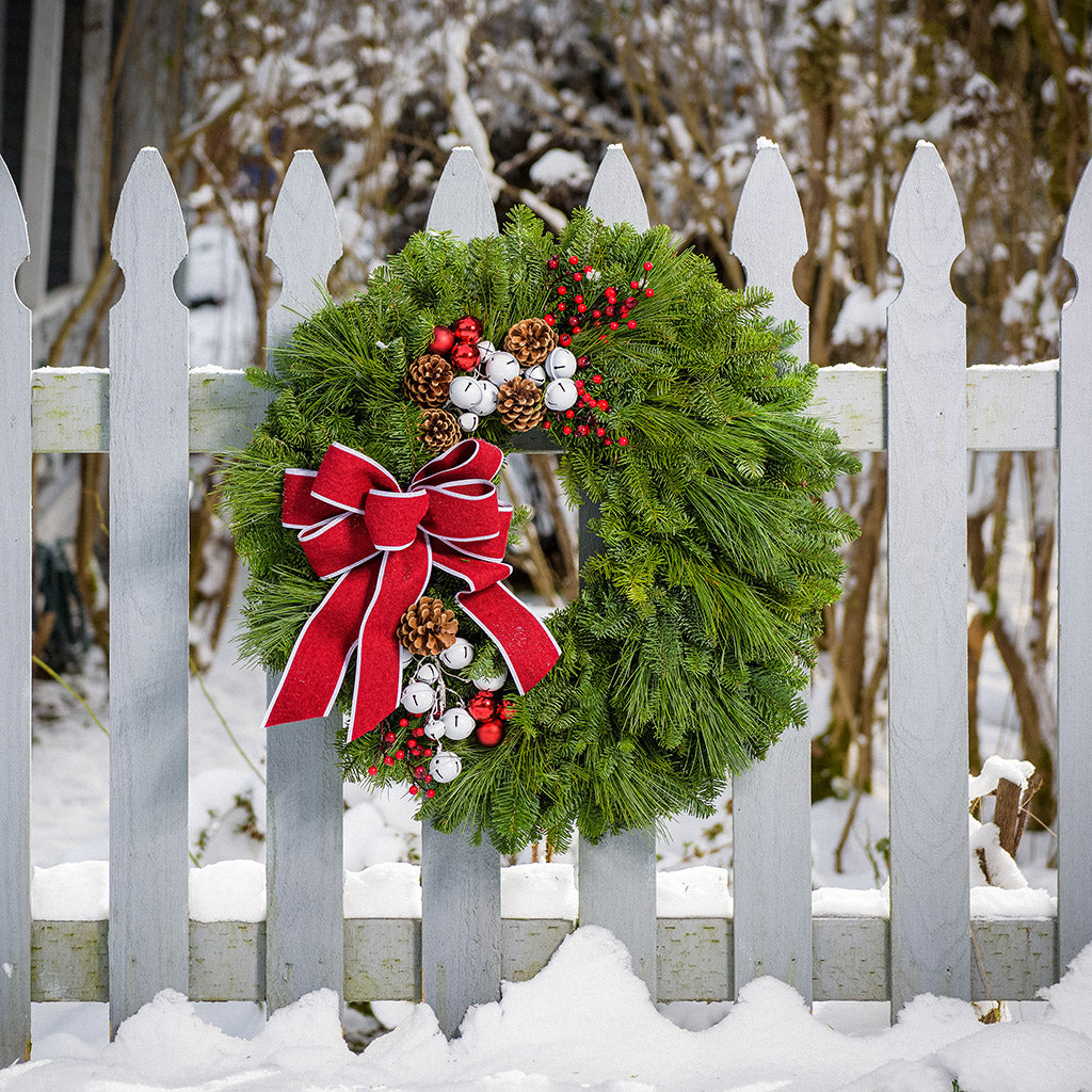 A holiday wreath made of noble fir and white pine with white jingle bells, 3 red berry clusters, 5 Australian pinecones, 2 red ball clusters, and a brushed red linen bow with white edging on a wood background.