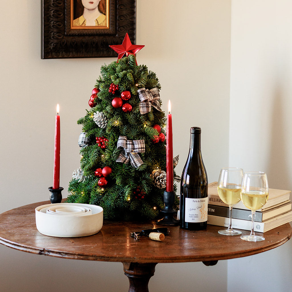 18” miniature tree decorated with white and black plaid bows, shiny red balls and berries, frosted pinecones and topped with a shiny red star tree-topper sitting on a wood round on a wooden table. 