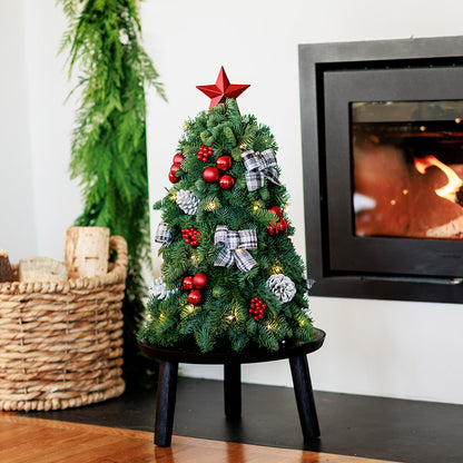 18” miniature tree decorated with white and black plaid bows, shiny red balls and berries, frosted pinecones and topped with a shiny red star tree-topper sitting on a wood round on a wooden table. 