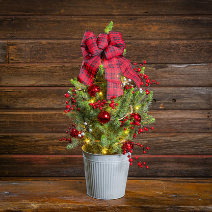 A small artificial Christmas tree in a galvanized metal bucket, decorated with a large red plaid bow, red berries, red ornaments, and warm white string lights, set against a dark wood plank background.