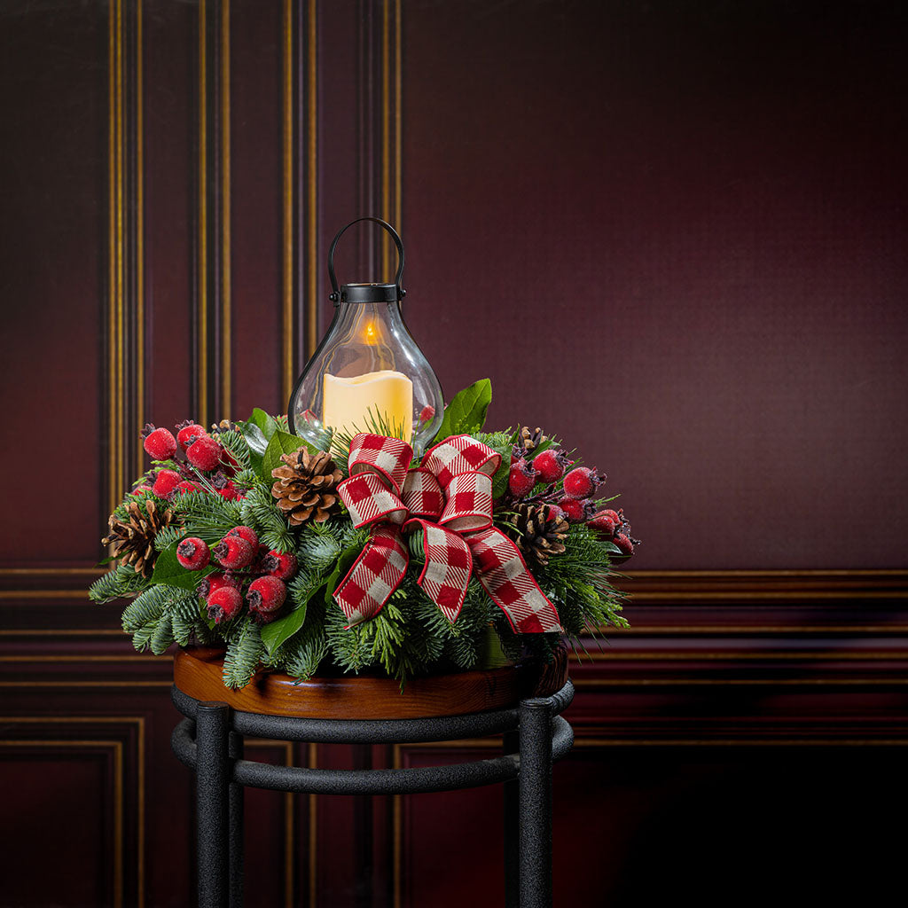 A festive holiday centerpiece arrangement on a black metal stand, featuring green pine branches, red berries, pinecones, a red-and-white gingham ribbon bow, and a lit glass lantern candle against a dark wood-paneled background.