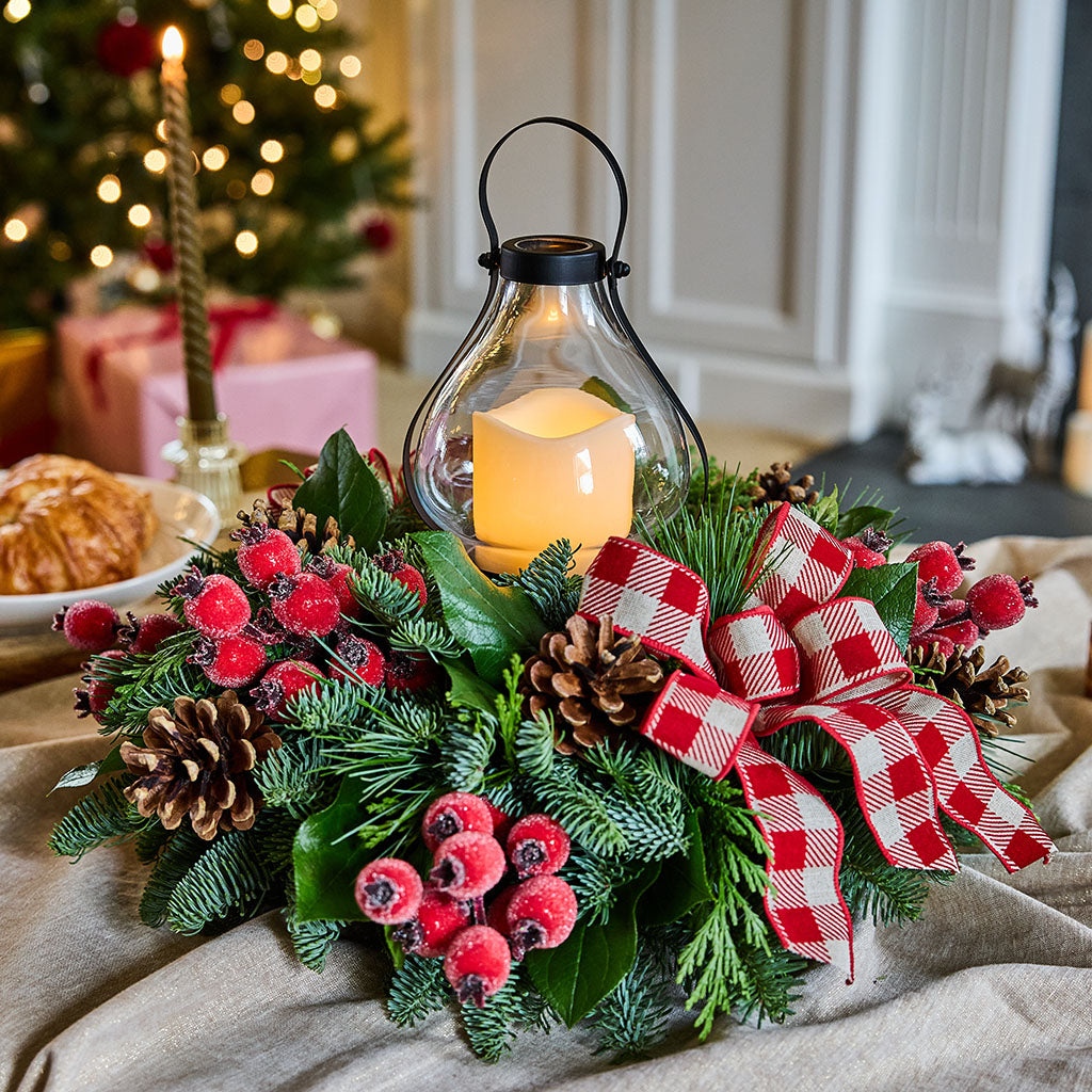 A festive Christmas centerpiece with green pine branches, red berry clusters, pinecones, a black metal lantern, and a red-and-white gingham ribbon bow, styled on a beige fabric surface with a blurred holiday background.