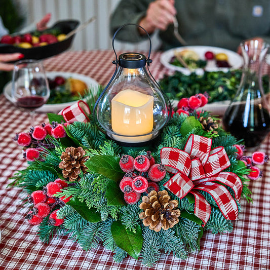 A festive holiday centerpiece on a red-and-white checkered tablecloth, featuring green pine branches, red berries, pinecones, a red gingham bow, and a lit glass lantern candle. Plates and a wine decanter are blurred in the background at a dinner table.