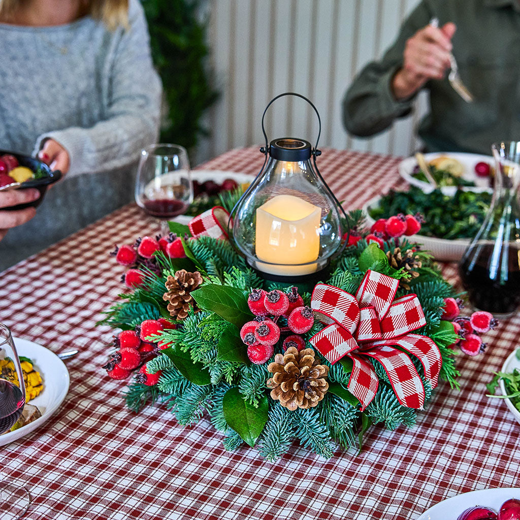 A festive Christmas wreath centerpiece on a red and white checkered tablecloth, featuring green pine branches, red berries, pinecones, a red gingham bow, and a lit glass lantern candle. Partial views of people dining are visible in the background.
