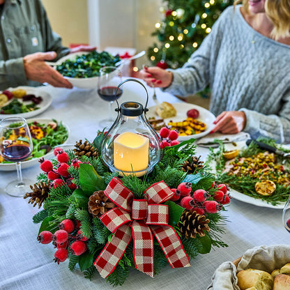 A festive Christmas centerpiece on a dining table, featuring a green pine wreath with red berries, pinecones, and a red-and-white gingham bow, with a glowing glass lantern candle in the center. Plates of food, wine glasses, and people seated at the table are visible in the background.