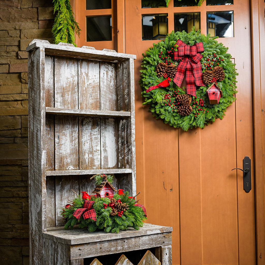 A holiday centerpiece of noble fir, white pine, incense cedar, natural pinecone, red berry & red ball clusters, 2 cardinal birds, natural stick clusters, a plaid birdhouse, and 2 tartan bows