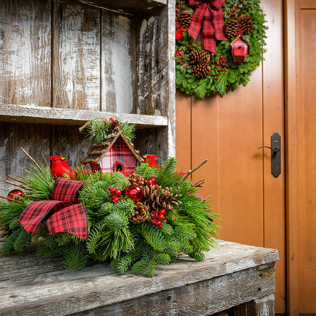 A holiday centerpiece of noble fir, white pine, incense cedar, natural pinecone, red berry & red ball clusters, 2 cardinal birds, natural stick clusters, a plaid birdhouse, and 2 tartan bows