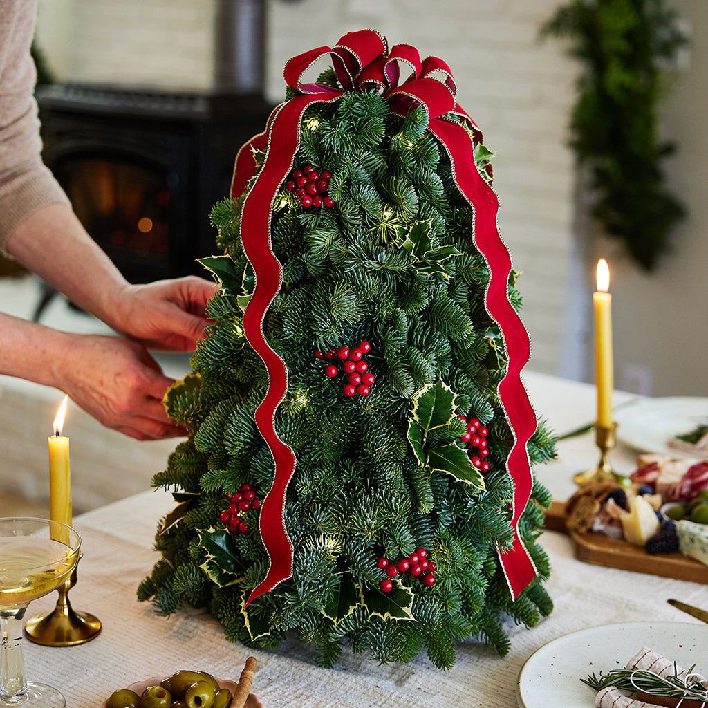 Fresh hand-assembled tabletop Christmas tree with fir, variegated holly, red berries and a red velveteen bow and white LEDs