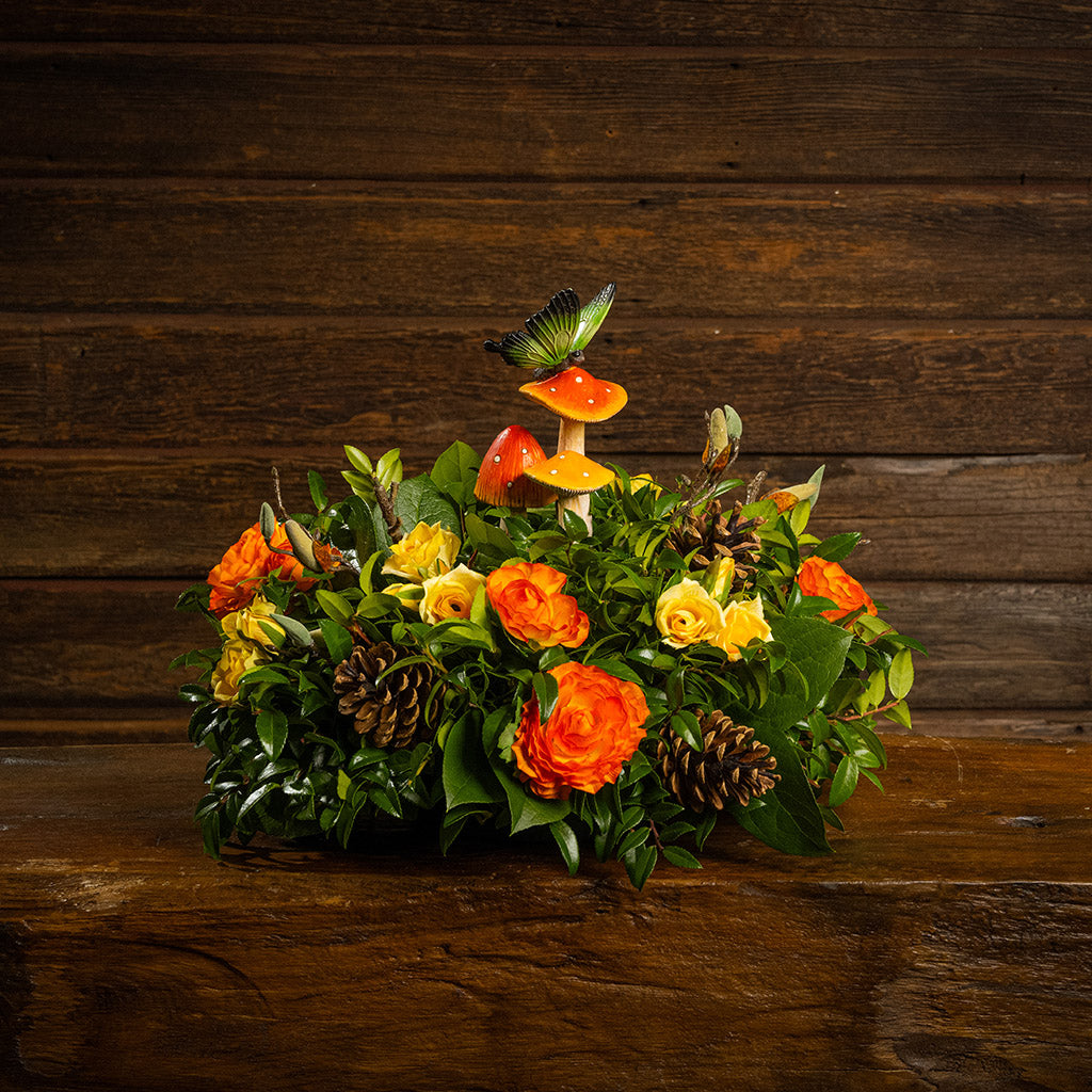 Fresh salal and green huckleberry with faux mini yellow roses, faux orange ranunculus flowers, faux magnolia branches, natural pinecones, and a resin mushroom & butterfly decoration