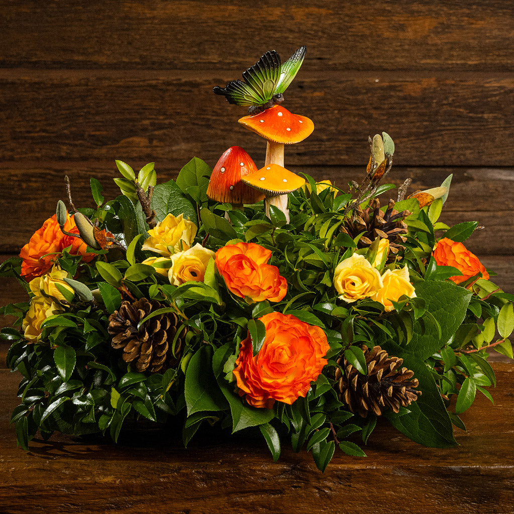 Fresh salal and green huckleberry with faux mini yellow roses, faux orange ranunculus flowers, faux magnolia branches, natural pinecones, and a resin mushroom & butterfly decoration