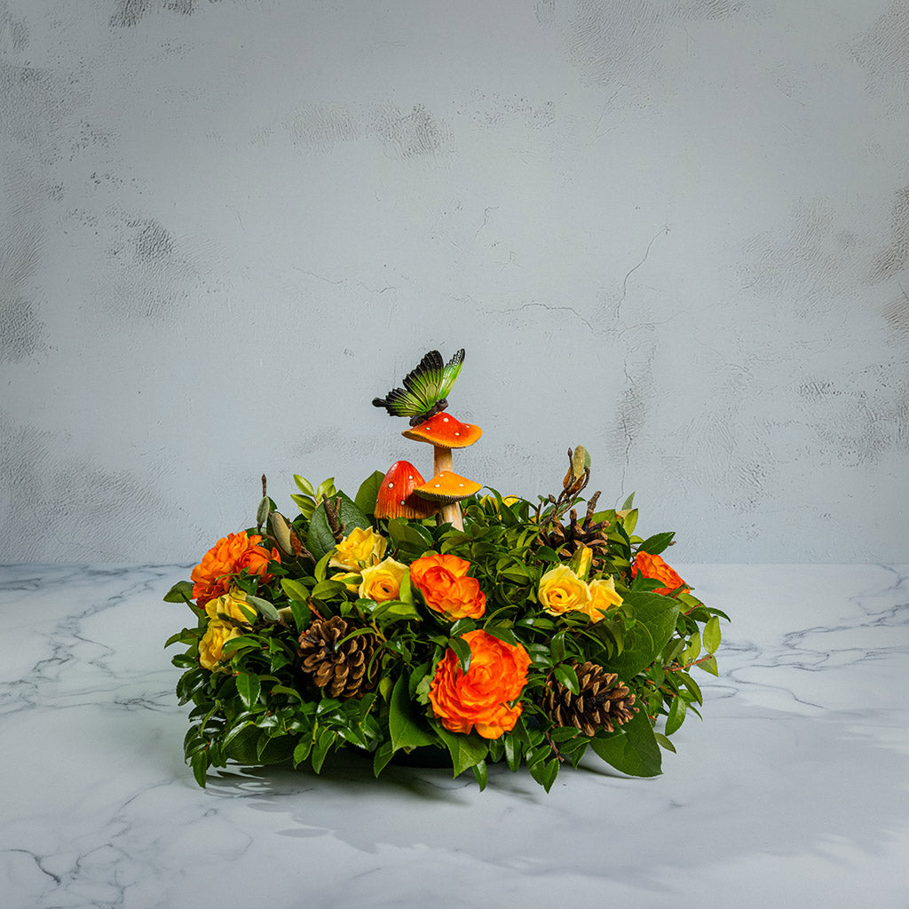 Fresh salal and green huckleberry with faux mini yellow roses, faux orange ranunculus flowers, faux magnolia branches, natural pinecones, and a resin mushroom & butterfly decoration