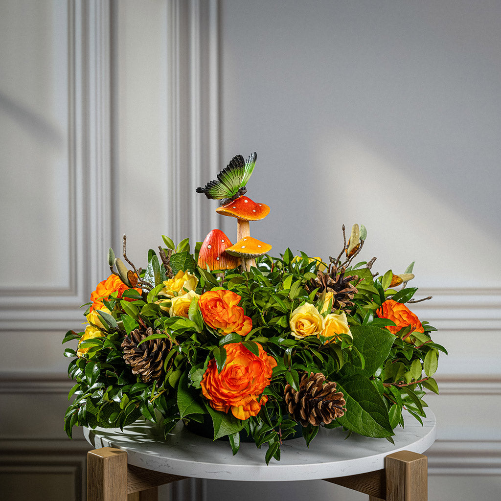 Fresh salal and green huckleberry with faux mini yellow roses, faux orange ranunculus flowers, faux magnolia branches, natural pinecones, and a resin mushroom & butterfly decoration