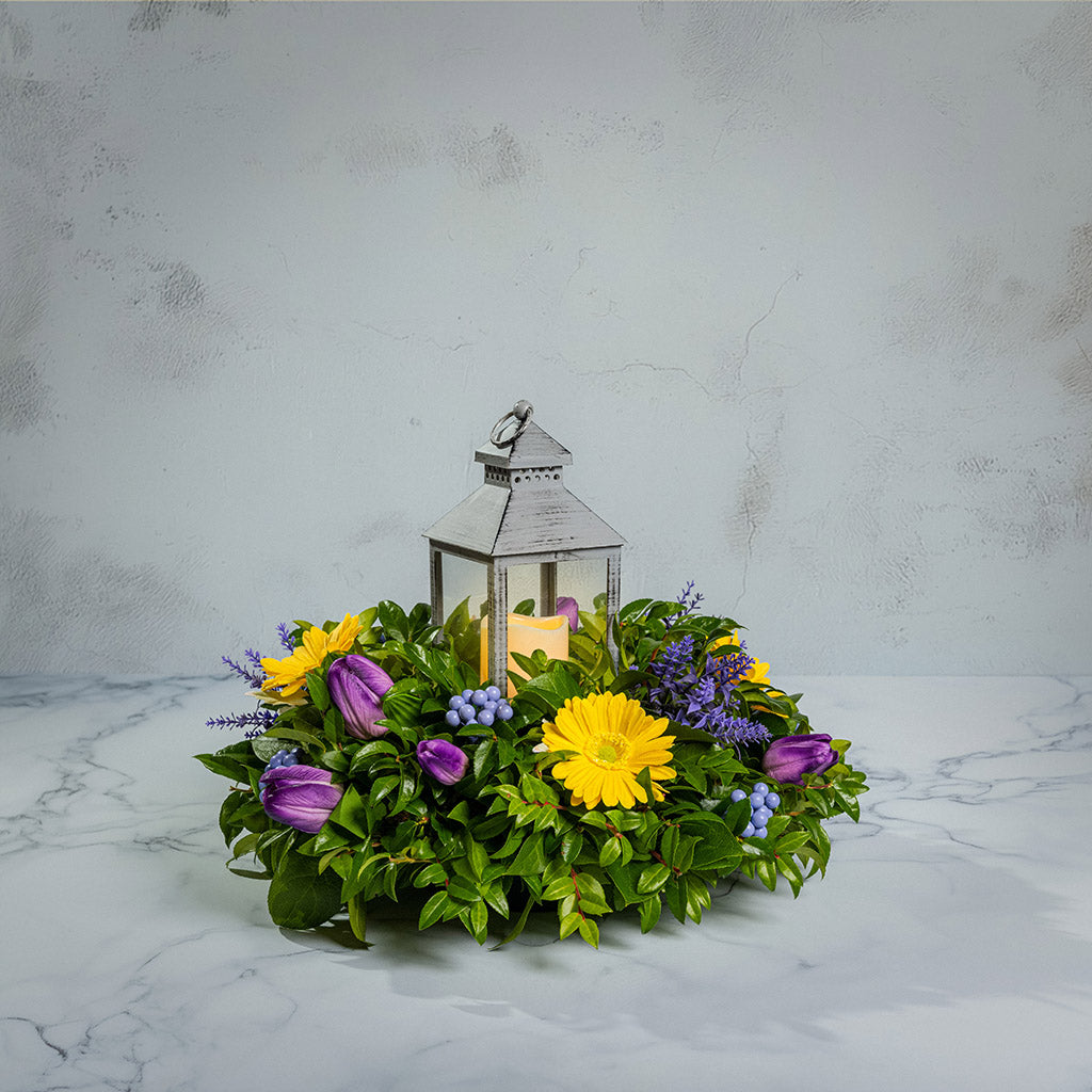 Fresh salal and green huckleberry with faux yellow gerbera daisies, faux purple tulips, faux lavender, and faux purple berries with a gray LED lantern