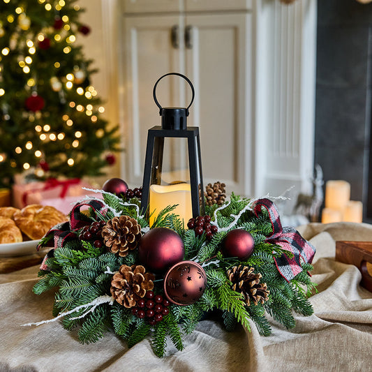 A festive Christmas wreath centerpiece on a draped beige fabric table runner, featuring green pine branches, brown pinecones, red ornaments, a black lantern with a lit candle, and red plaid ribbon accents. A blurred Christmas tree with warm lights is visible in the background.