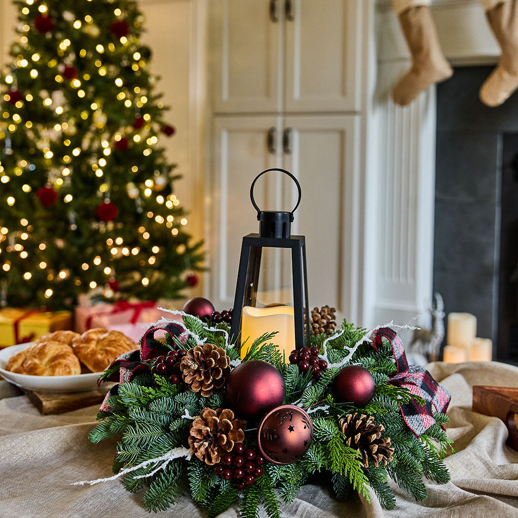 A festive Christmas wreath centerpiece on a table, featuring green pine branches, dark red ornaments, pinecones, a black lantern with a lit candle, and a plaid ribbon, set in a cozy holiday kitchen with a blurred Christmas tree in the background.