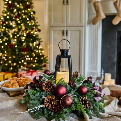 A festive Christmas wreath centerpiece on a table, featuring green pine branches, dark red ornaments, pinecones, a black lantern with a lit candle, and a plaid ribbon, set in a cozy holiday kitchen with a blurred Christmas tree in the background.