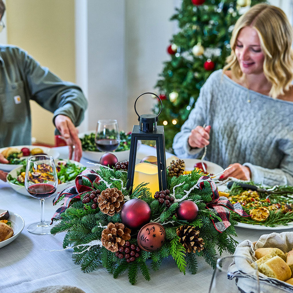 A festive Christmas table centerpiece with pine branches, red ornaments, pinecones, and a black lantern candle, set on a dining table with people seated at a Christmas tree in the background.