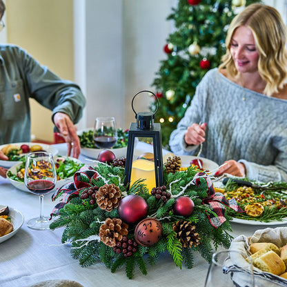 A festive Christmas table centerpiece with pine branches, red ornaments, pinecones, and a black lantern candle, set on a dining table with people seated at a Christmas tree in the background.