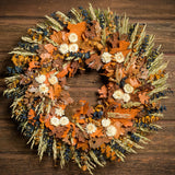 A close-up of a dried wreath made of preserved spiral eucalyptus in amber and navy blue colors with a mix of sudan grass, preserved oak leaves in orange and cafe colors, and cream strawflower accents hung on a dark wood background.