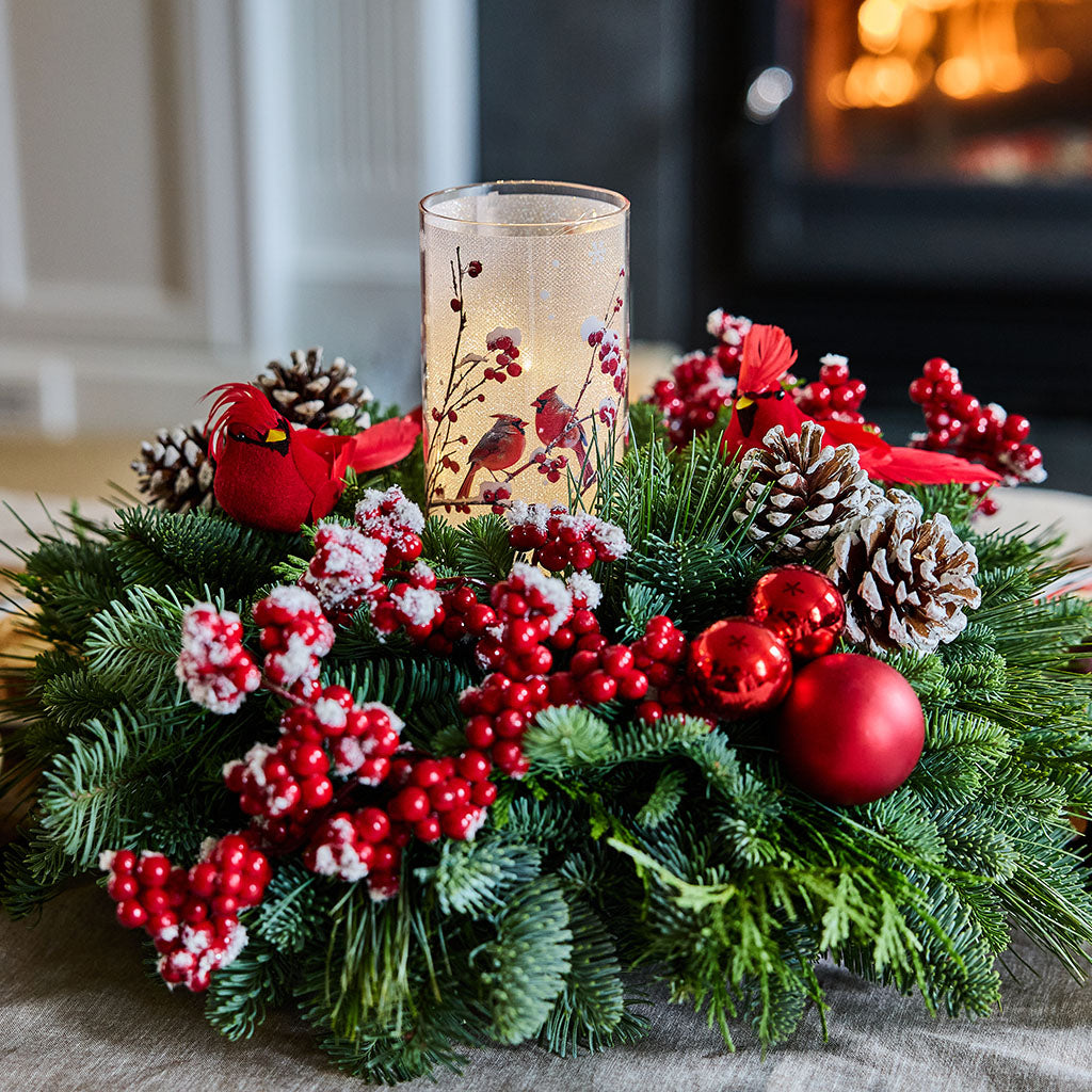 A festive Christmas wreath made of green pine branches, decorated with red berries, white snow-dusted accents, pinecones, red ornaments, and two red cardinal figurines, with a clear glass candle holder featuring a bird design placed in the center. The wreath is set on a neutral fabric surface with a blurred indoor background showing a warm fireplace.