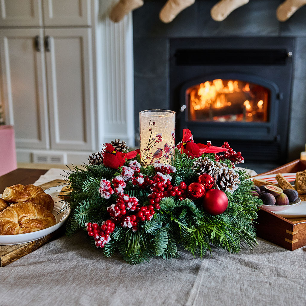A festive Christmas wreath centerpiece on a table in a cozy living room, featuring green pine branches, red berries, pinecones, red ornaments, and a lit candle, with a fireplace in the background.