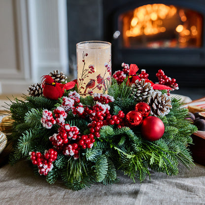 A festive Christmas wreath made of green pine branches, decorated with red berries, pinecones, red ornaments, and a lit glass candle holder, set on a table in front of a warm fireplace.