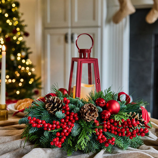 A festive Christmas wreath centerpiece on a draped tablecloth, featuring green pine and fir branches, red berries, pinecones, and a red lantern, with a blurred holiday tree and fireplace in the background.