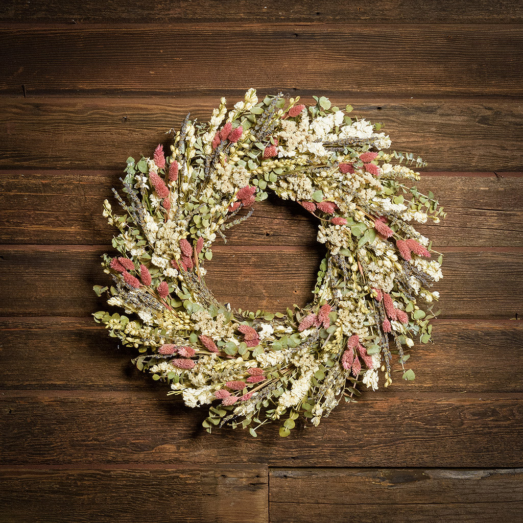 A dried wreath made of natural English lavender, sage green eucalyptus, white statice blooms and pink phalaris hung on a dark wood background.