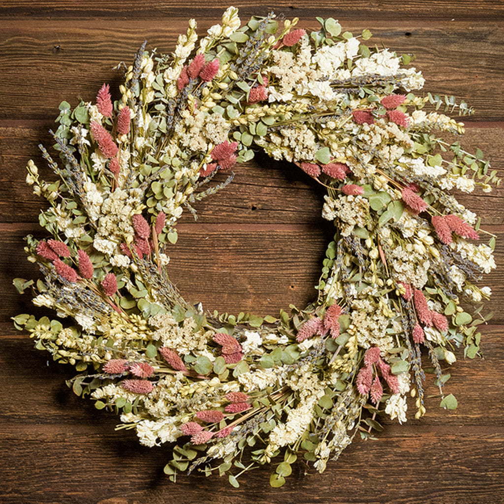 A close-up of a dried wreath made of natural English lavender, sage green eucalyptus, white statice blooms and pink phalaris hung on a dark wood background.