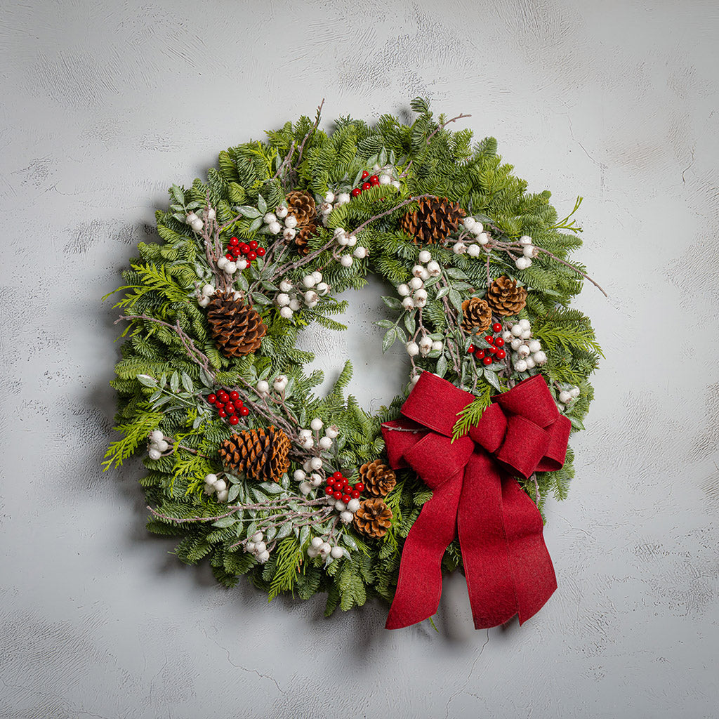 Christmas wreath with greenery, pinecones, berries, and a red bow on a light gray background