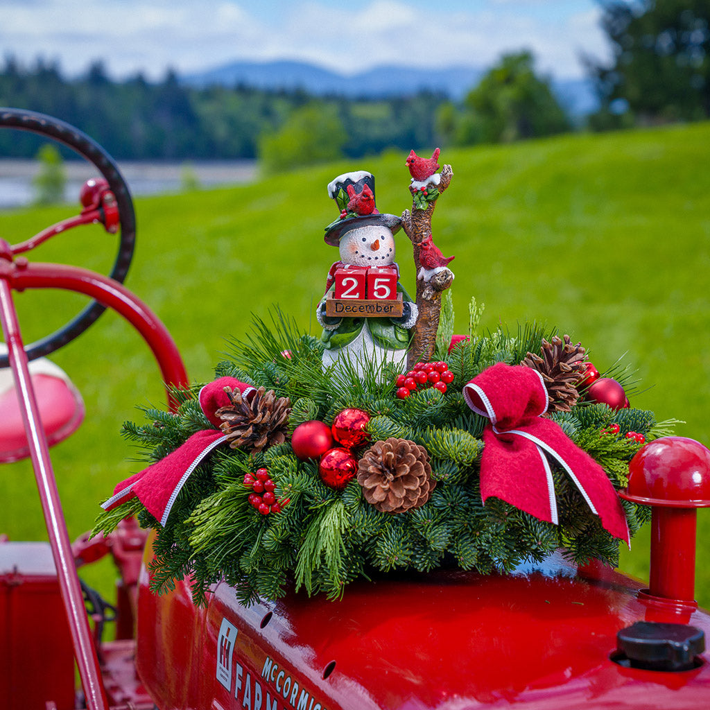 A centerpiece of noble fir, cedar, and pine with red ball ornaments, faux red berries, pinecones, 3 red bow tucks, and a decoration of a resin snowman holding 2 red blocks with numbers representing the number of days left until Christmas. Next to the black-top hat-wearing snowman is a frosted tree with 2 red cardinals sitting on a red tractor outside.